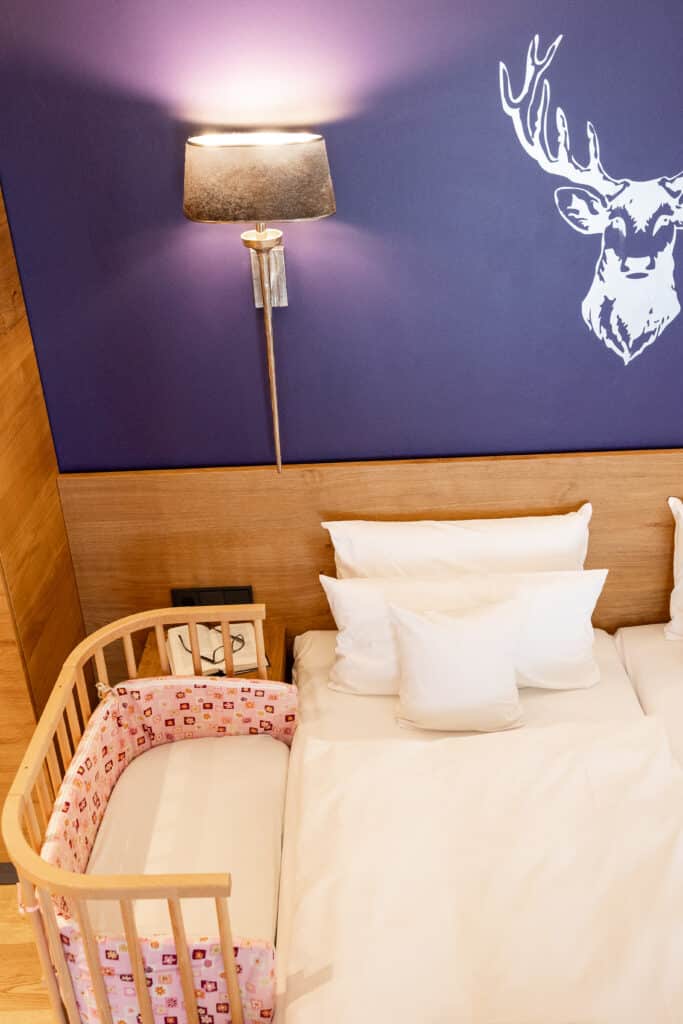 Baby-Beistellbett neben Elternbett vor einer lilanen Wand mit silbernem Hirschkopf