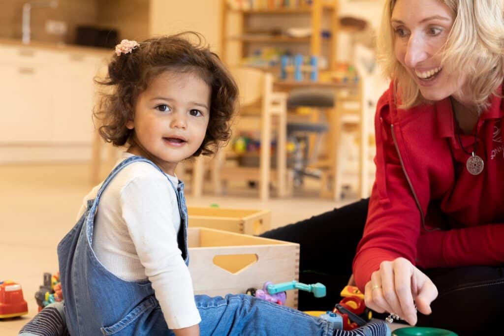 Blonde Frau lächelt einem kleinen Mädchen in blauer Latzhose zu vor einer Babybetreuung