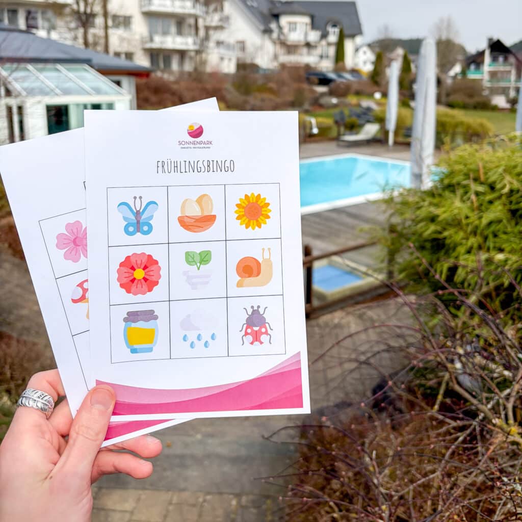 Hand hält zwei Frühlingsbingo-Karten aufgefächert in die Höhe vor einem Pool im Hintergrund