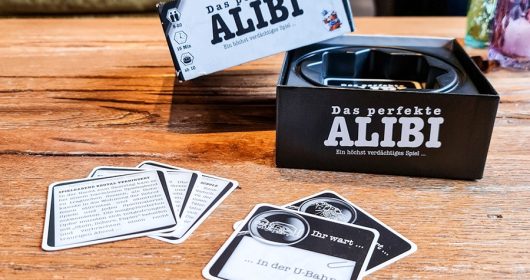 Spiel "Das perfekte Alibi" auf einem Holztisch mit Spielkarten davor