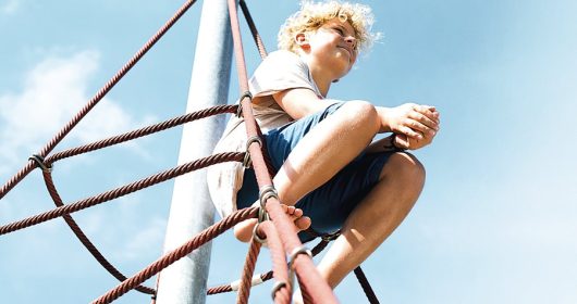 Junge sitzt Barfuß auf einem Klettegerüst bei blauem Himmel und schaut zufrieden in die Ferne