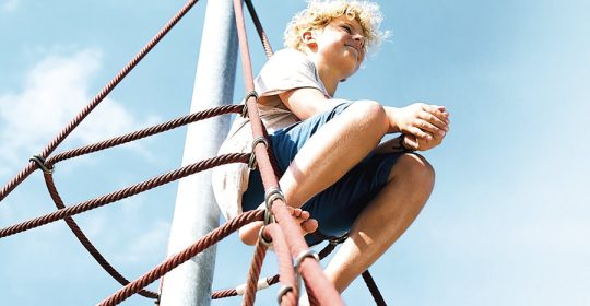 Junge sitzt Barfuß auf einem Klettegerüst bei blauem Himmel und schaut zufrieden in die Ferne