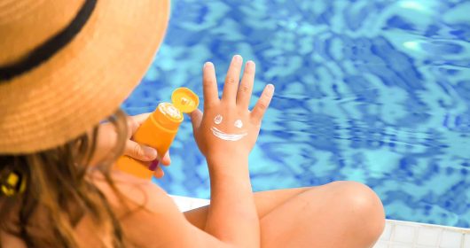 Mädchen mit Strohhut sitzt am Pool und schmiert sich mit Sonnencreme einen Smiley auf die Hand.
