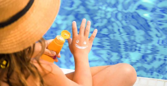 Mädchen mit Strohhut sitzt am Pool und schmiert sich mit Sonnencreme einen Smiley auf die Hand.