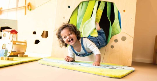 Kleines Mädchen in blauer Latzhose krabbelt aus hölzernem Spieltunnel mit grünen Vorhängen