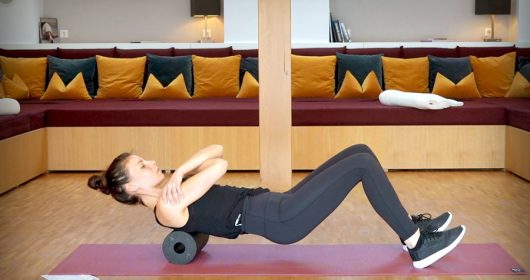Frau absolviert eine Übung auf einer schwarzen Faszienrolle vor einem großen Sofa