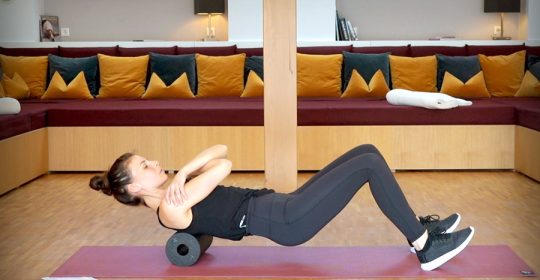 Frau absolviert eine Übung auf einer schwarzen Faszienrolle vor einem großen Sofa