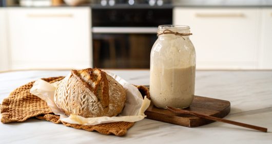 Gebackenes Brot auf weißem Tuch neben einem Glas mit Sauerteiganstellgut vor einer Küche