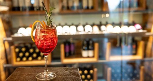 Ein Glas Aperol Winter Spritz auf einer Holztheke vor einem Weinregal in der Lounge vom Familotel Sonnenpark