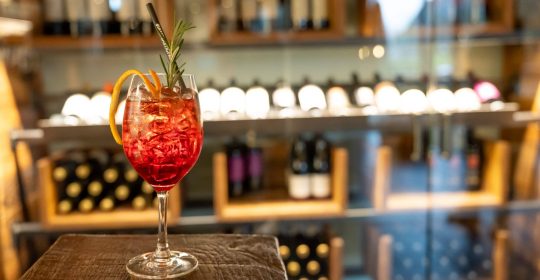 Ein Glas Aperol Winter Spritz auf einer Holztheke vor einem Weinregal in der Lounge vom Familotel Sonnenpark