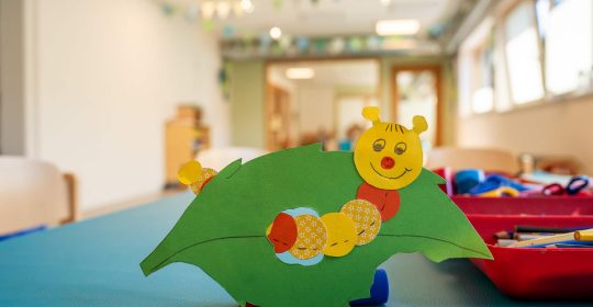 Bunte Raupe auf grünem Blatt aus Tonkarton auf einem Basteltisch mit Bastelutensilien