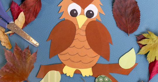 Gebastelte braune Eule aus Tonkarton umgeben von Herbstlaub und Bastelutensilien auf einer blauen Tischdecke
