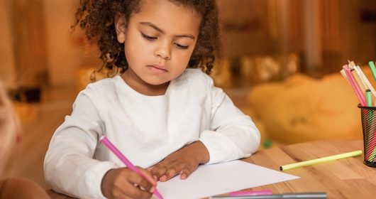 Kind mit schwarzen Locken malt etwas auf weißem Papier an einem Holztisch