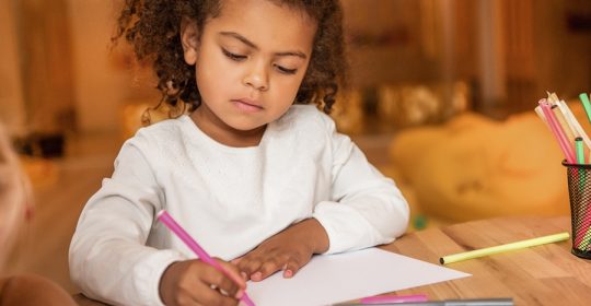 Kind mit schwarzen Locken malt etwas auf weißem Papier an einem Holztisch