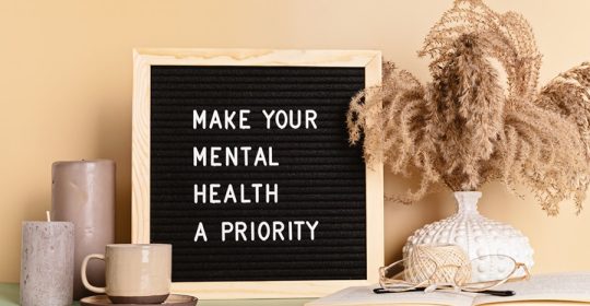 "Machen Sie Ihre geistige Gesundheit zu einer Priorität" Motivationszitat auf dem Bilderrahmen, umgeben von Dekoelementen wie Kerzen, Tasse, Buch