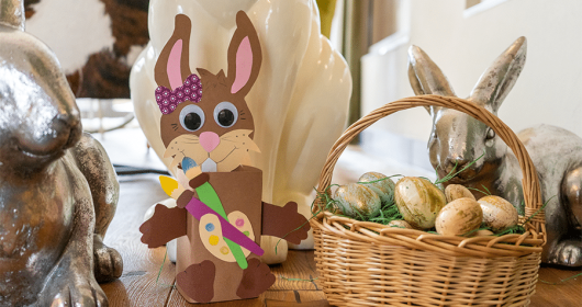 Bonboniere in Form eines Osterhasens neben einem gefüllten Osterkörbchen, umgeben von Dekohasen