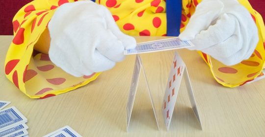 Clown mit weißen Handschuhen versucht ein Kartenhaus aufzubauen