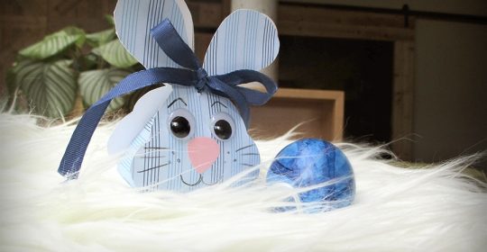 blaue Tonkartonbox mit Hasengesicht neben einem blauen Osterei auf Schafsfell