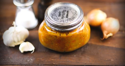 Glas mit Auberginenaufstrich vor Gewürzen, Zwiebeln und Knoblauch auf einem Holztisch