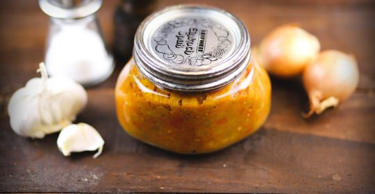Glas mit Auberginenaufstrich vor Gewürzen, Zwiebeln und Knoblauch auf einem Holztisch