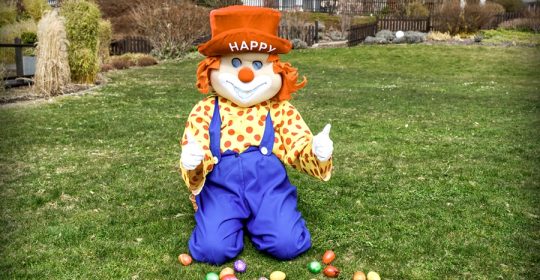 Clown "Happy" sitzt auf einer grünen Wiese vor bunten Ostereiern