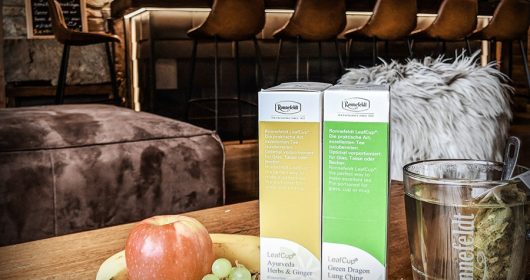 Teeverpackungen und Teeglas neben Obst auf einem Holztisch in einer Lounge