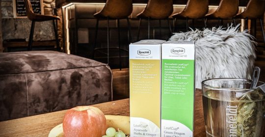 Teeverpackungen und Teeglas neben Obst auf einem Holztisch in einer Lounge