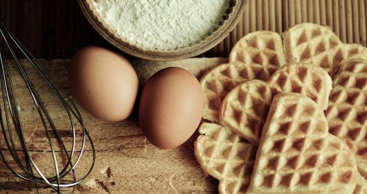 Waffeln neben Eiern, Mehl und einem Rührstab auf einem Holzbrett