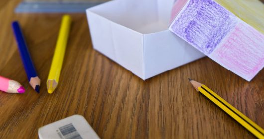 Buntstifte und Radiergummi vor einer Papierkiste auf Holztisch