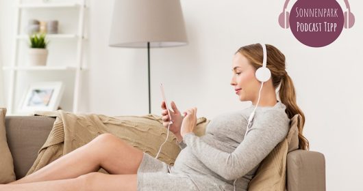 Schwangere Frau in grauem Kleid liegt auf dem Sofa mit Kopfhörern und hört einen Podcast