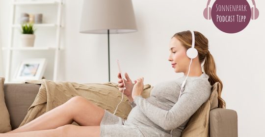 Schwangere Frau in grauem Kleid liegt auf dem Sofa mit Kopfhörern und hört einen Podcast