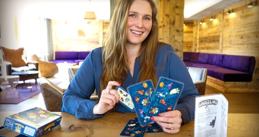 Frau in blauer Bluse präsentiert Spielkarten von "Gruselino" in einer Lounge