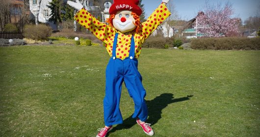 Clown "Happy" mit rotem Hut hüpft bei Sonnenschein auf dem Rasen