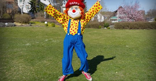 Clown "Happy" mit rotem Hut hüpft bei Sonnenschein auf dem Rasen