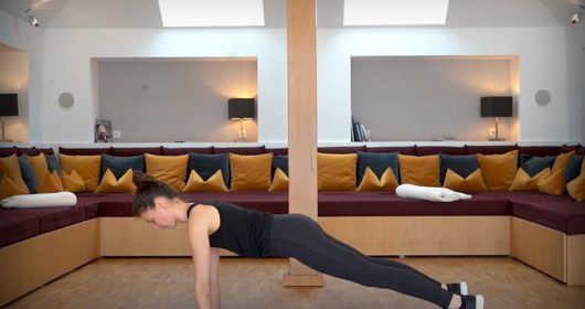 Frau in dunklem Sportoutfit macht eine Yoga-Pose auf einer rosa Matte vor einem Sofa mit vielen Kissen