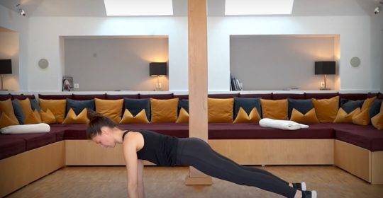 Frau in dunklem Sportoutfit macht eine Yoga-Pose auf einer rosa Matte vor einem Sofa mit vielen Kissen