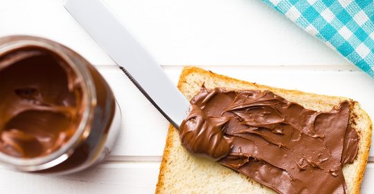 Eine Toastscheibe wird mit Schokocreme bestrichen auf einem weißen Tisch