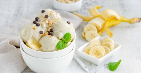 Selbstgemachtes veganes Bananeneis in einer Schüssel mit Erdnüssen und Schokolade vor einem hölzernen Hintergrund. Gesundes Dessert. Querformat, Freiraum