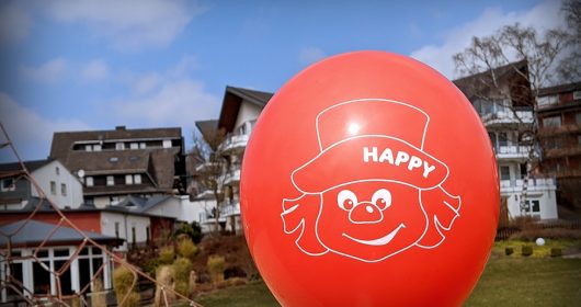 roter Ballon mit Clownsgesicht vor einem Haus auf einer grünen Wiese