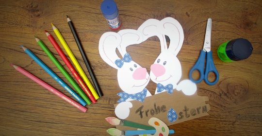 Zwei weiße HAsen aus Tonkarton vor einem Osterschild neben Bastelutensilien auf einem braunen Tisch