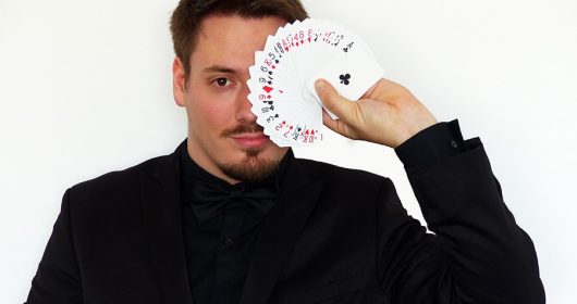 Mann in schwarzer Kleidung hält ein aufgefächertes Kartenspiel vor sein linkes Auge